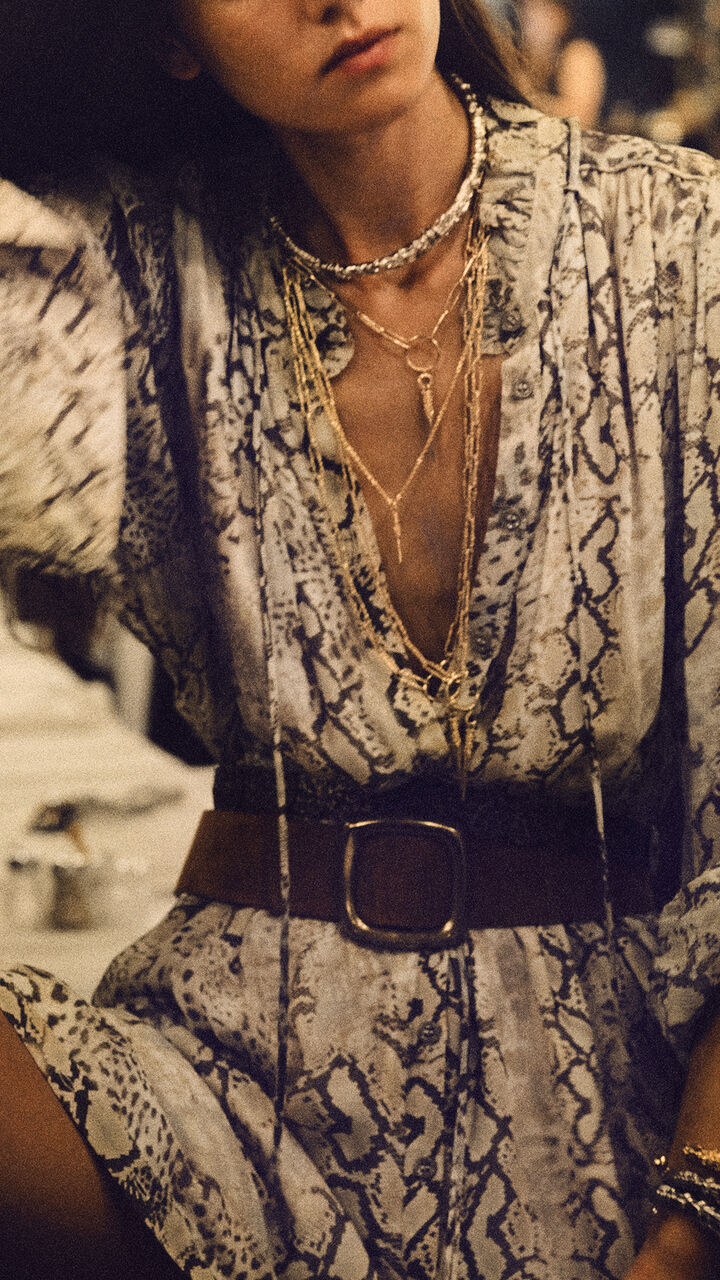 The model is wearing a long printed dress with a belt and several necklaces.