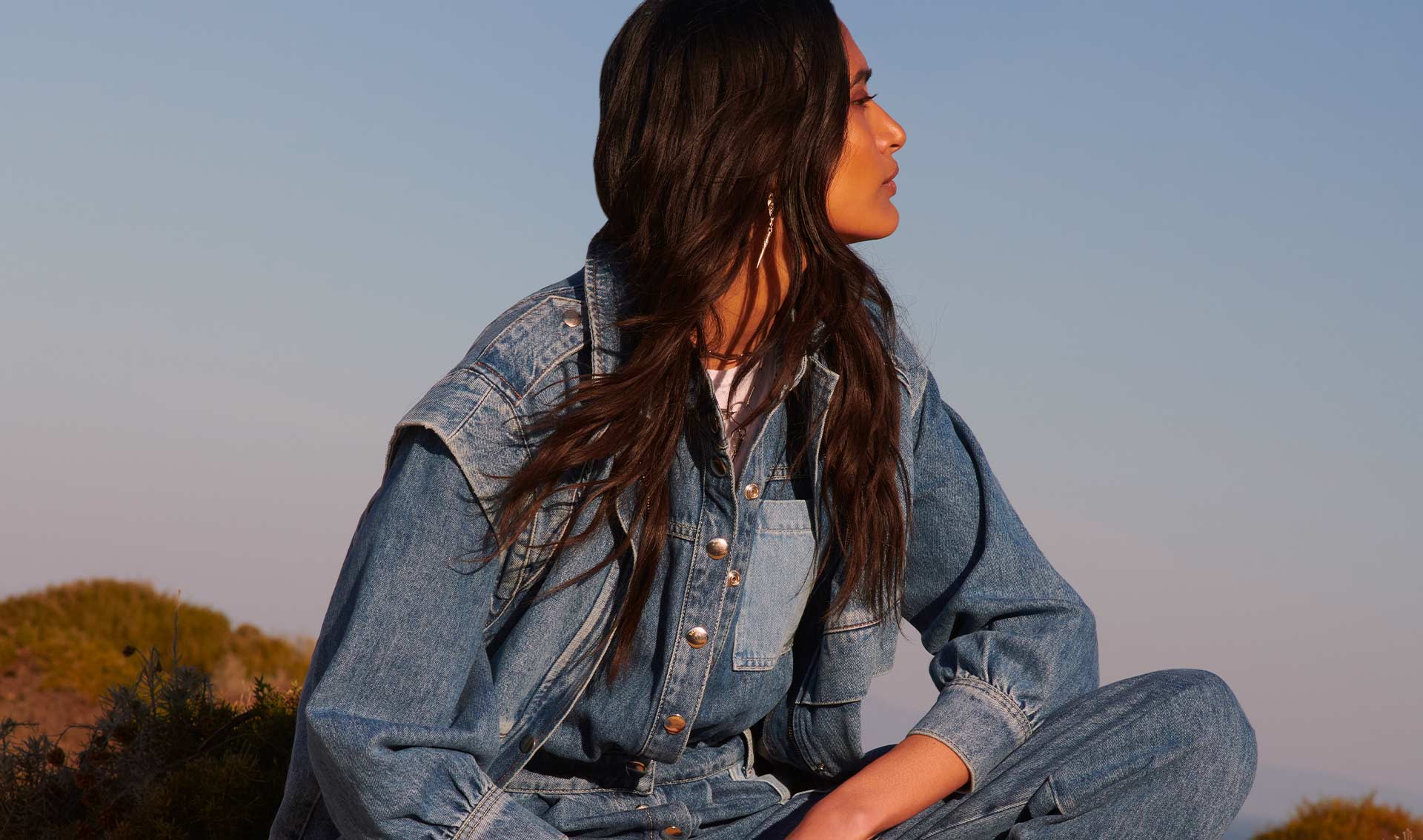 a brown-haired woman wearing a denim patchwork jumpsuit is looking at the horizon