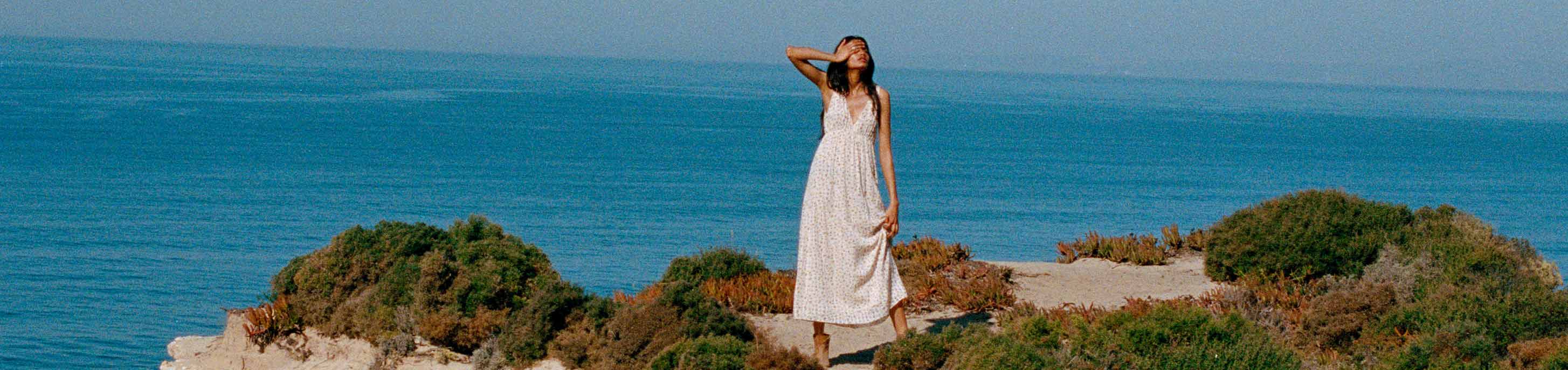 a girl standing on a cliff looking out into the ocean in a ba&sh long printed white maxi dress