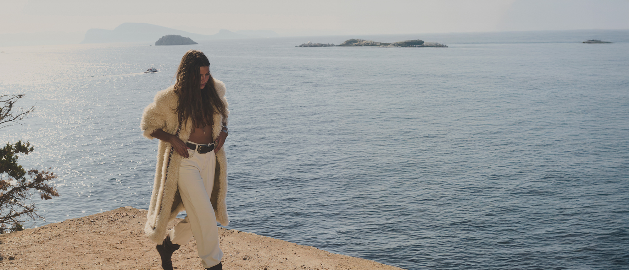 a model wearing a white fur coat, brown ankle boots, and white pants at the edge of a cliff overlooking the sea
