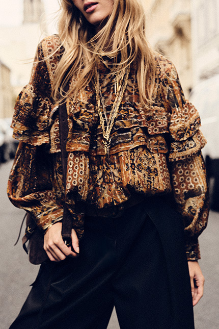 The model is wearing a patterned blouse and several necklaces