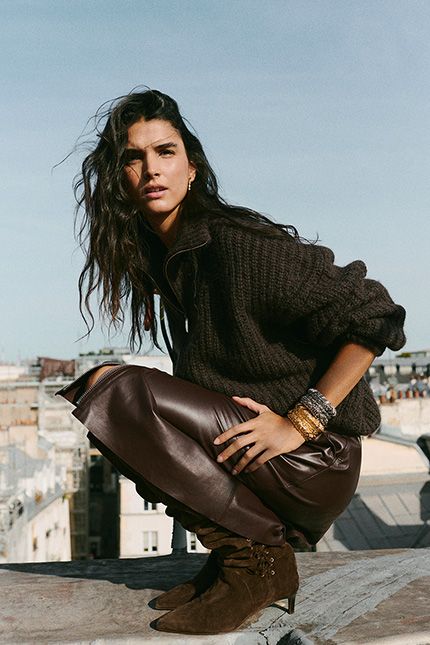 The model is wearing a brown knit sweater, a leather skirt, and boots 