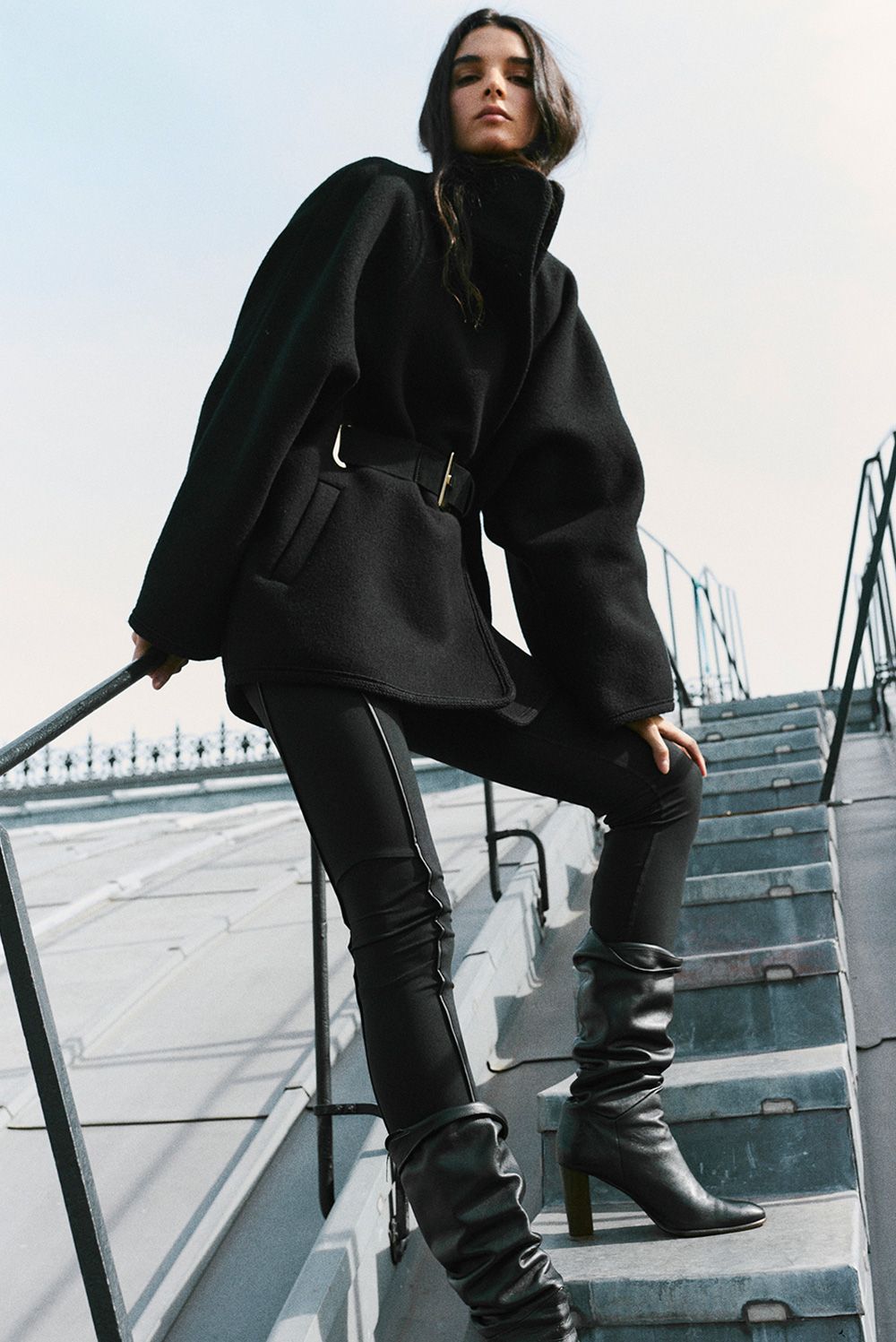 The model is wearing a black belted coat, jeans, and leather boots