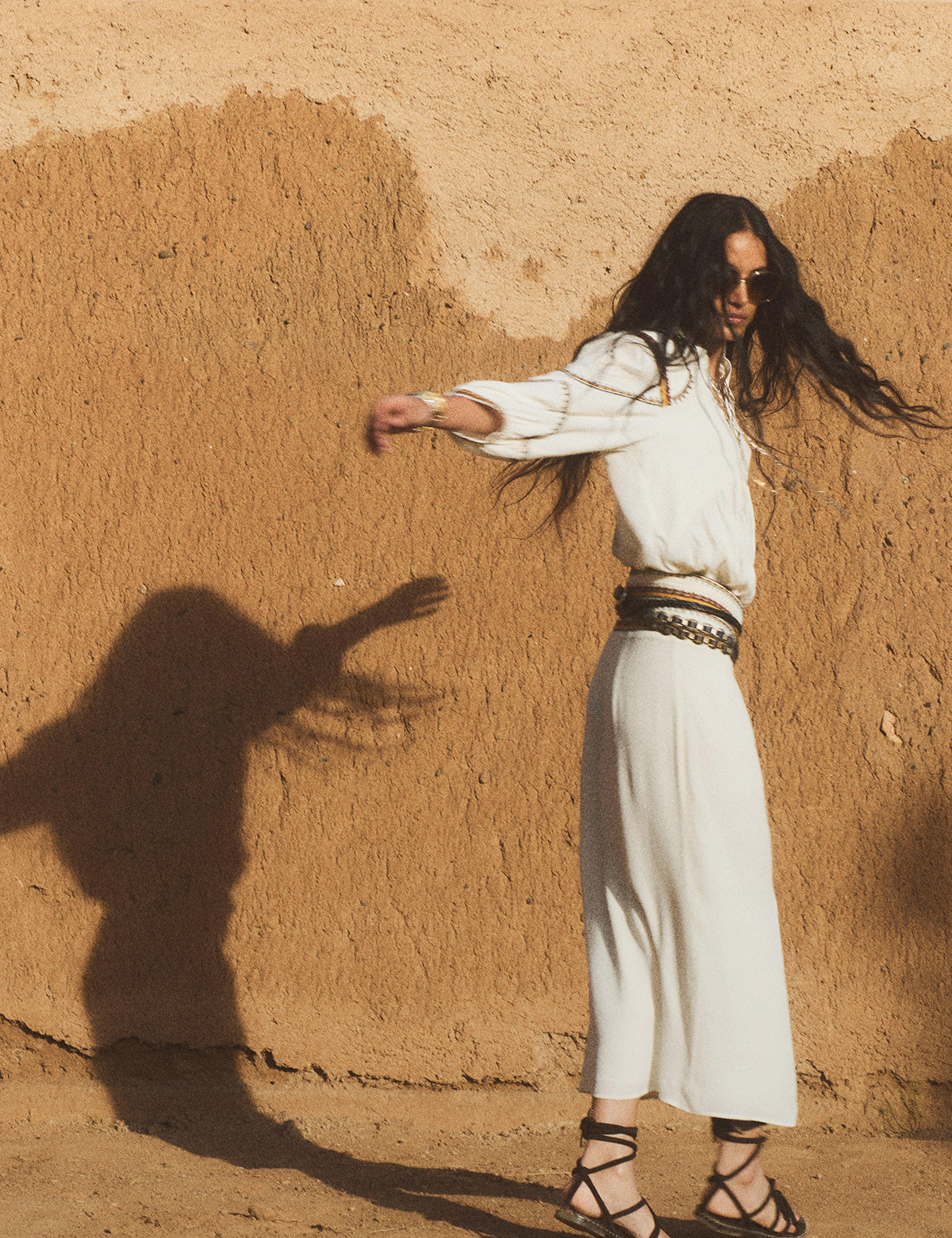 A model wearing a long white dress with several belts. She is standing in front of an orange-yellow wall.