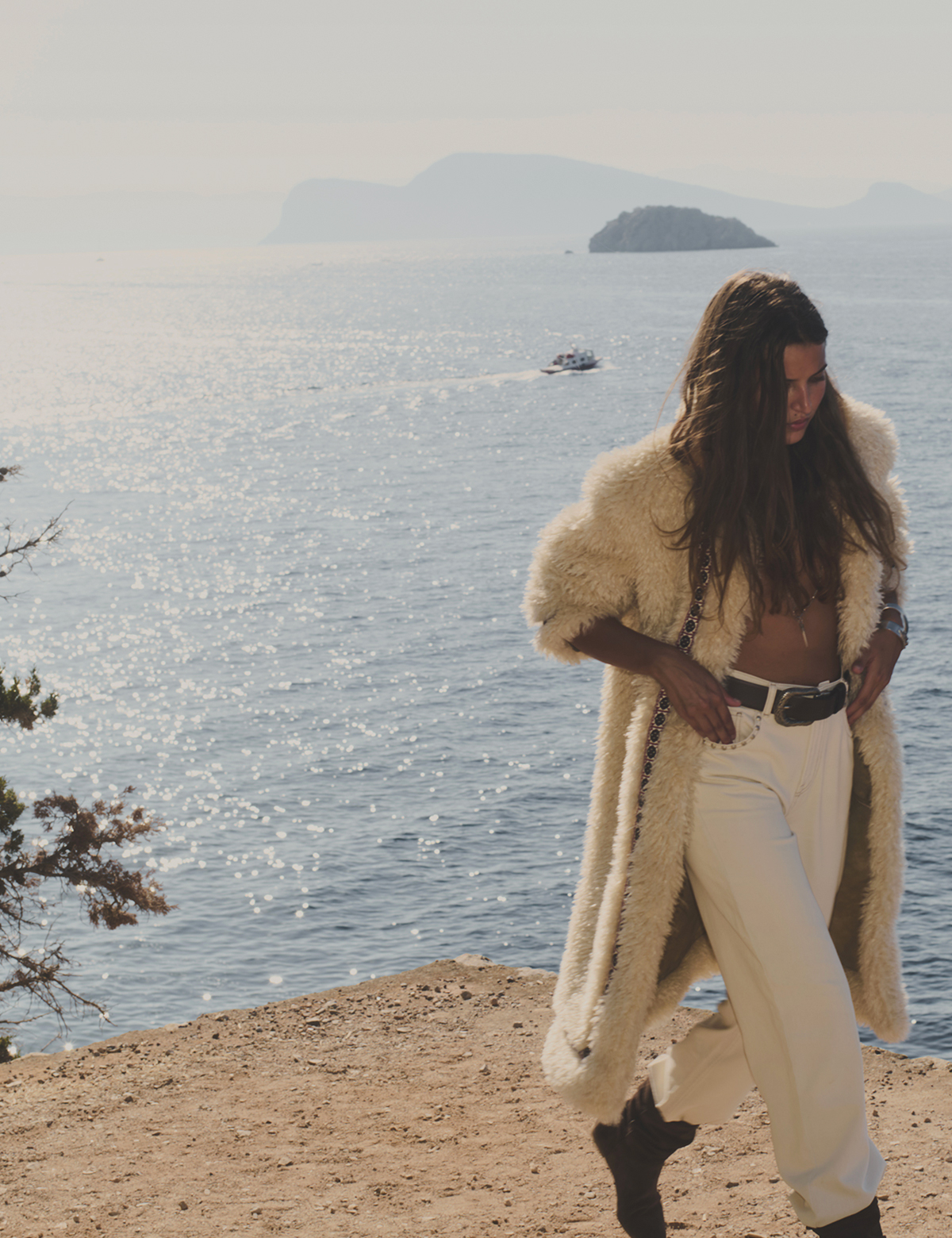 a model wearing a white fur coat, brown ankle boots, and white pants at the edge of a cliff overlooking the sea