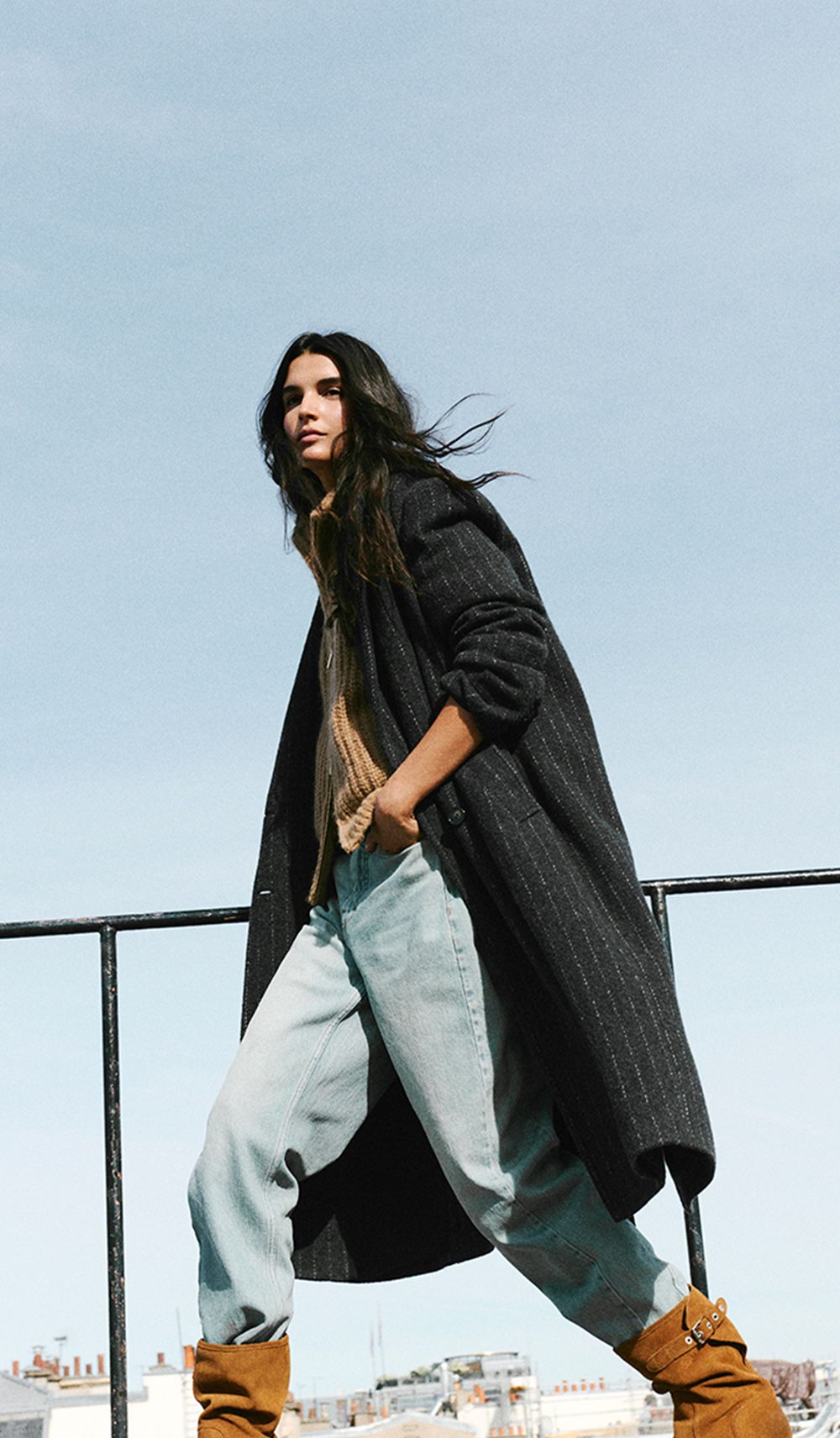 The model is wearing a long coat, a knit sweater, jeans, and suede boots