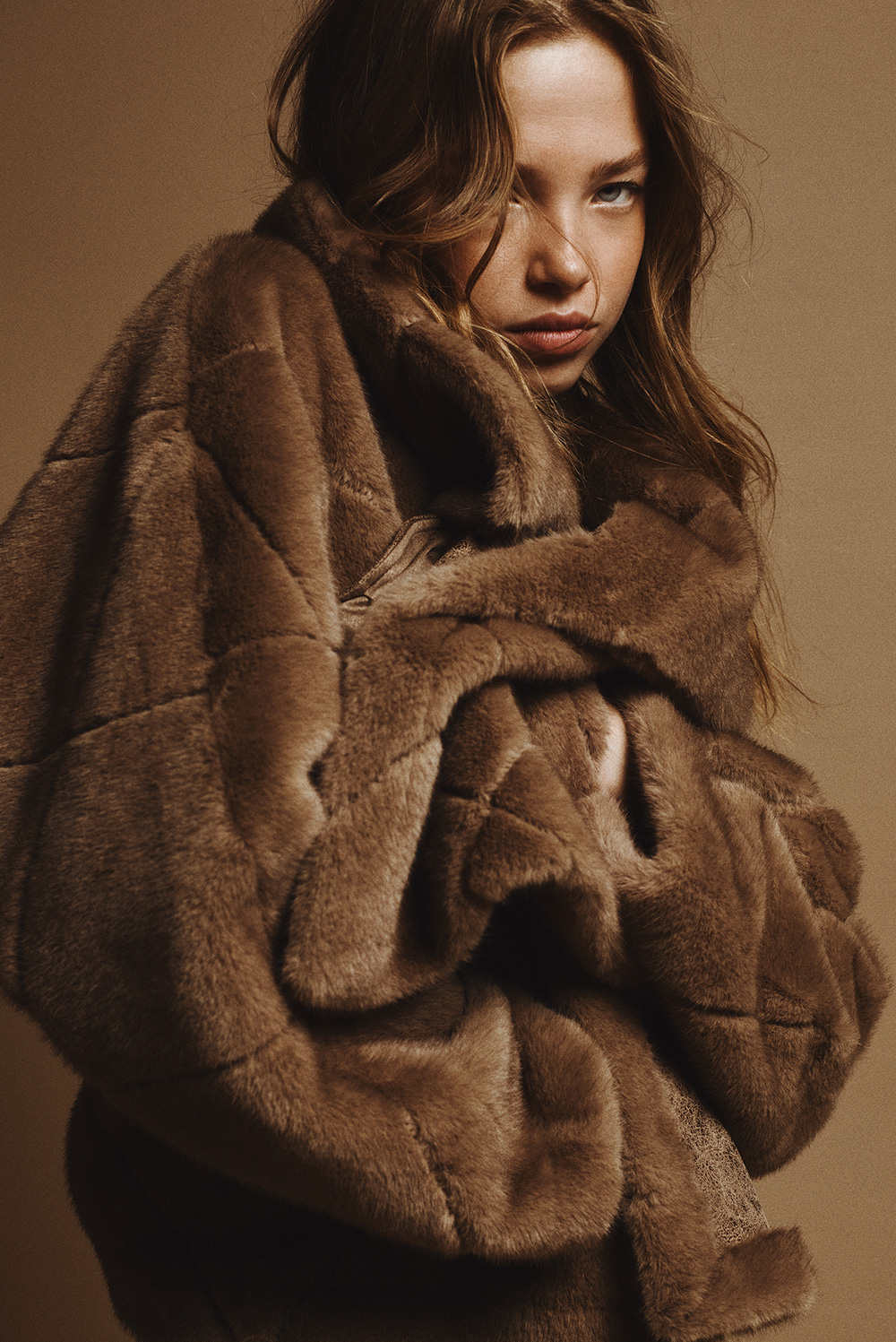 Model wearing an oversized brown faux fur coat styled with white jeans and suede boots, posing against a beige background — elegant winter fashion look.