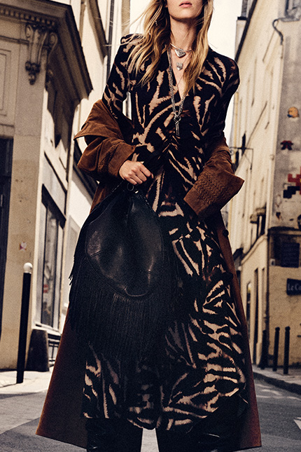 The model is wearing a long printed dress, a suede jacket, and a leather bag