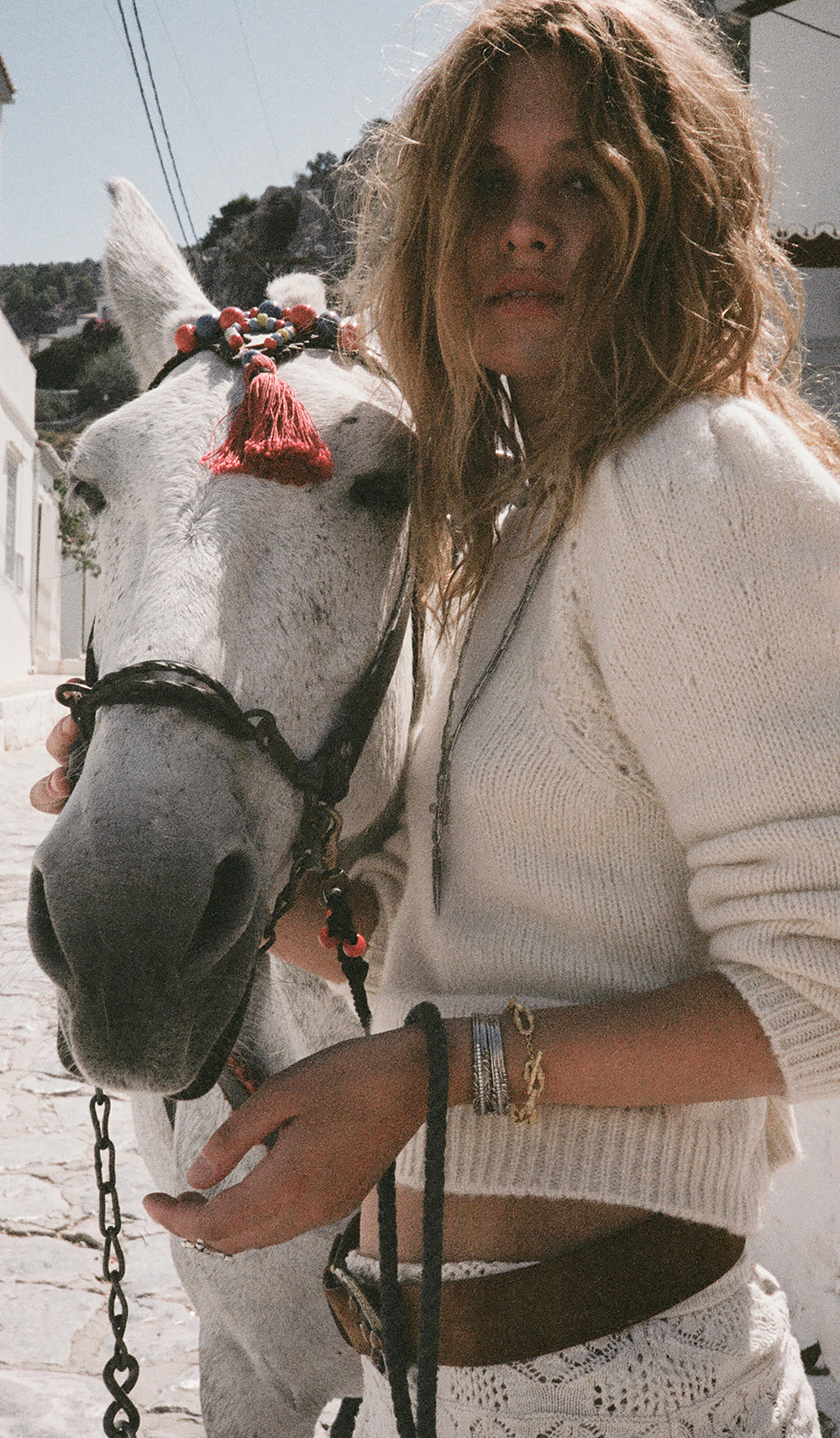 A blonde model wearing a white knit outfit pulling a white horse down a street 