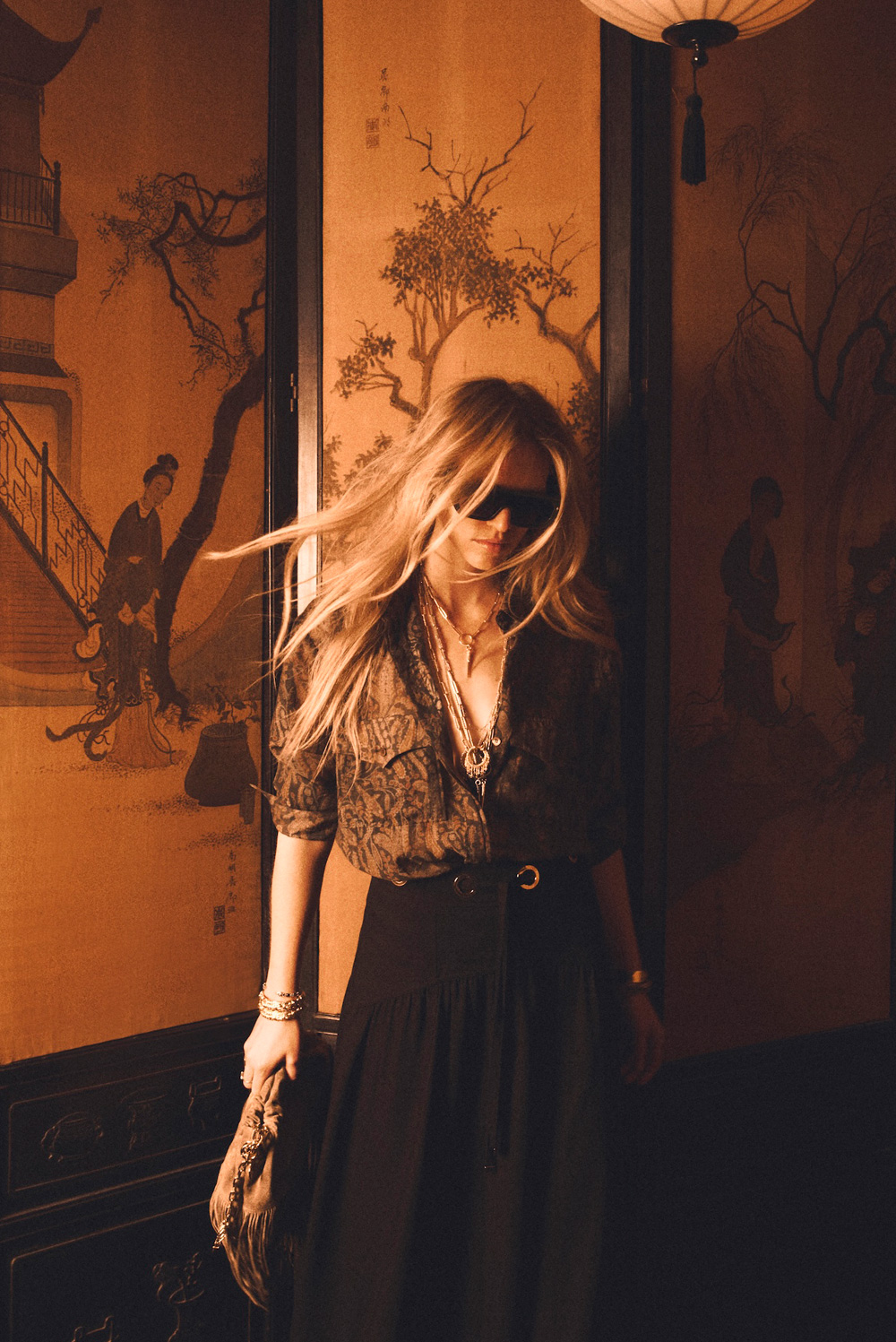 A blonde model wearing a printed shirt with flowing black trousers and sunglasses.