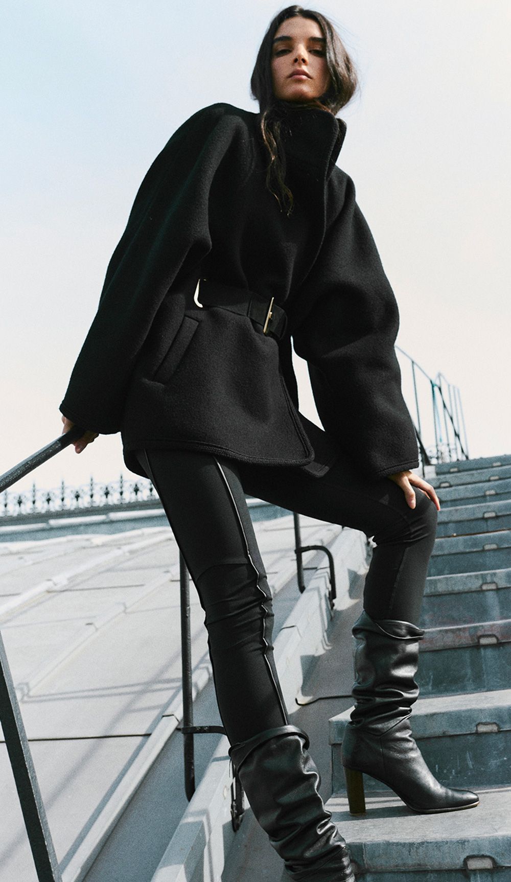 The model is wearing a black belted coat, jeans, and leather boots