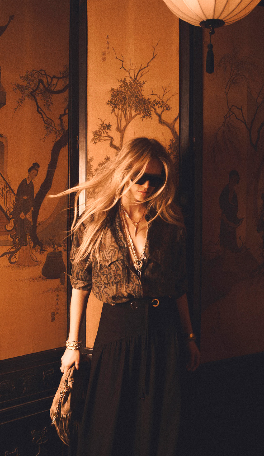 A blonde model wearing a printed shirt with flowing black trousers and sunglasses.